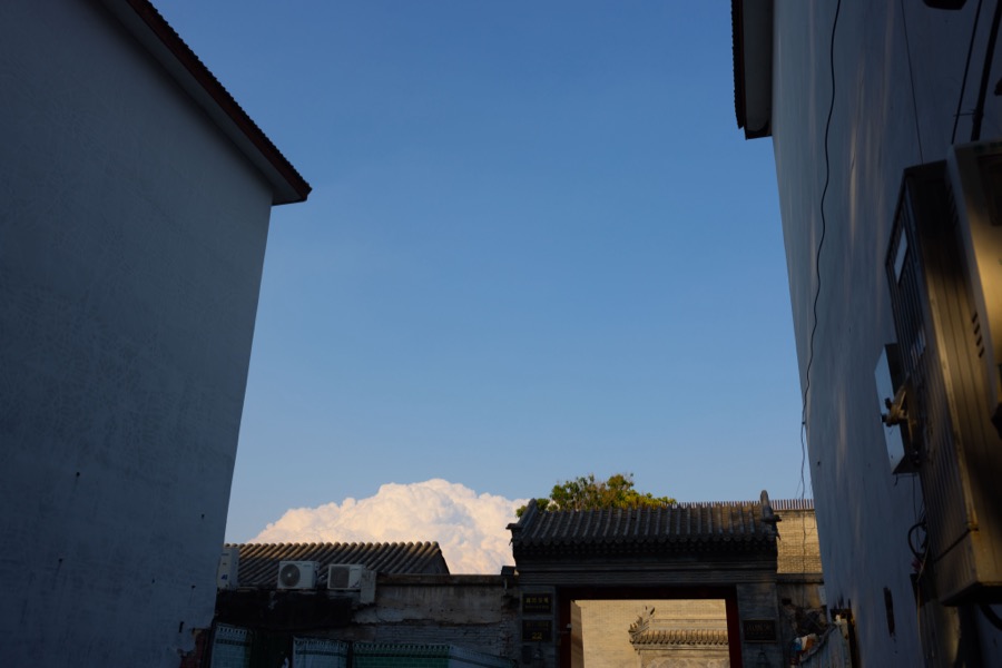 A narrow alley between two pale walls opens onto blue sky, with a bank of cloud rising above a tiled gateway.