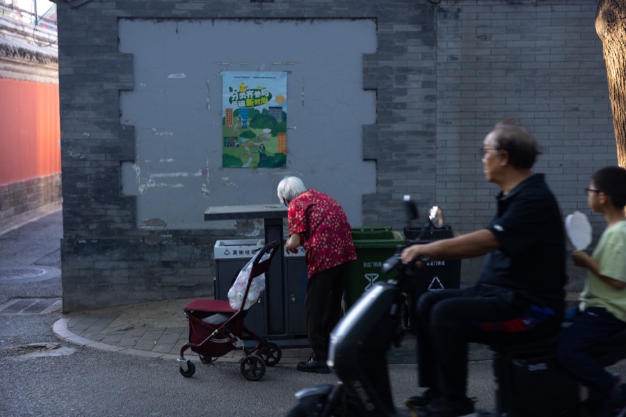 An elderly woman in a red floral shirt sorts through recycling bins by a grey brick wall as a man and a boy ride past on a scooter.