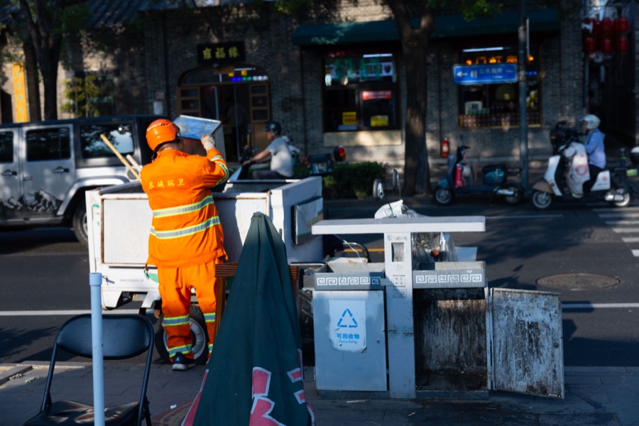 A sanitation worker in an orange uniform shields his eyes beside a cart and street bins in late sun.