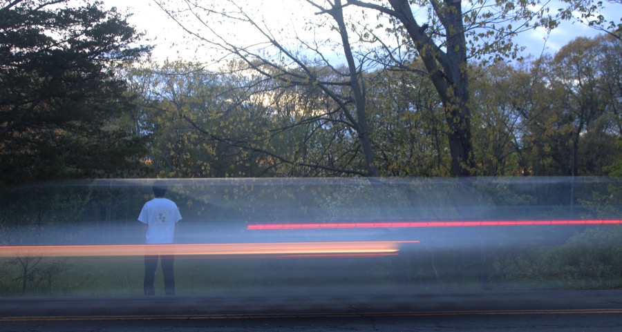A figure stands at the roadside as red and orange light trails streak past.