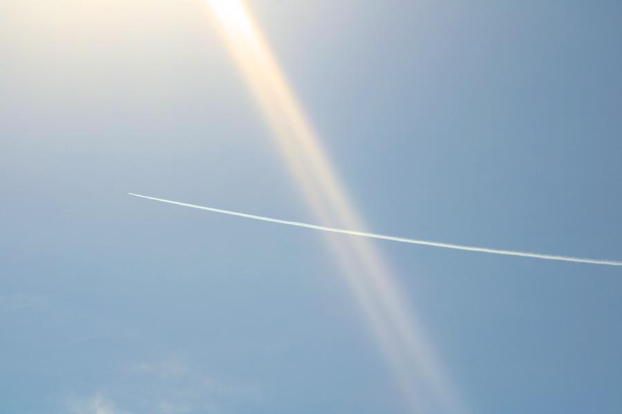 A thin contrail crosses a pale blue sky through a beam of sun flare.